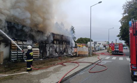 Obraz na wolnym powietrzu, ogień, niebo, strażak gasi pożar, dużo dymu, dwa samochody strażackie stoją na ulicy.