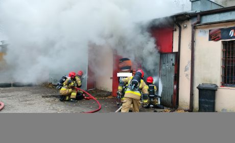 na zdjęciu widać czterech strażaków, którzy gaszą pożar. Strażacy znajdują się na zewnątrz budynku. Widać dużo dymu, który wydostaje się z budynku. Na ziemi leżą węże strażackie.