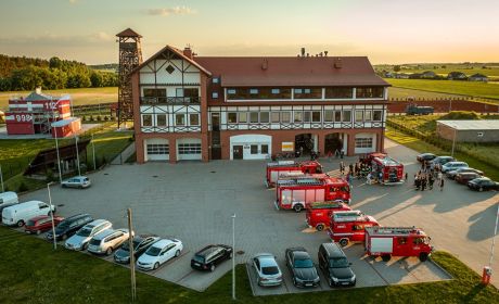 Na zdjęciu widać budynek, w oddali wieżę, drzewa, pola. Przed budynkiem znajduje się parking, na którym zaparkowane są samochody, zarówo straży pożarnej, jak i osobowe. Na parkingu stoją strażacy.