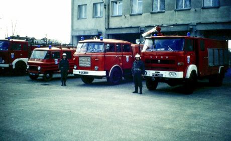 Przed budynkiem stoją trzy samochody strażackie. Obok nich stoi dwóch strażaków.